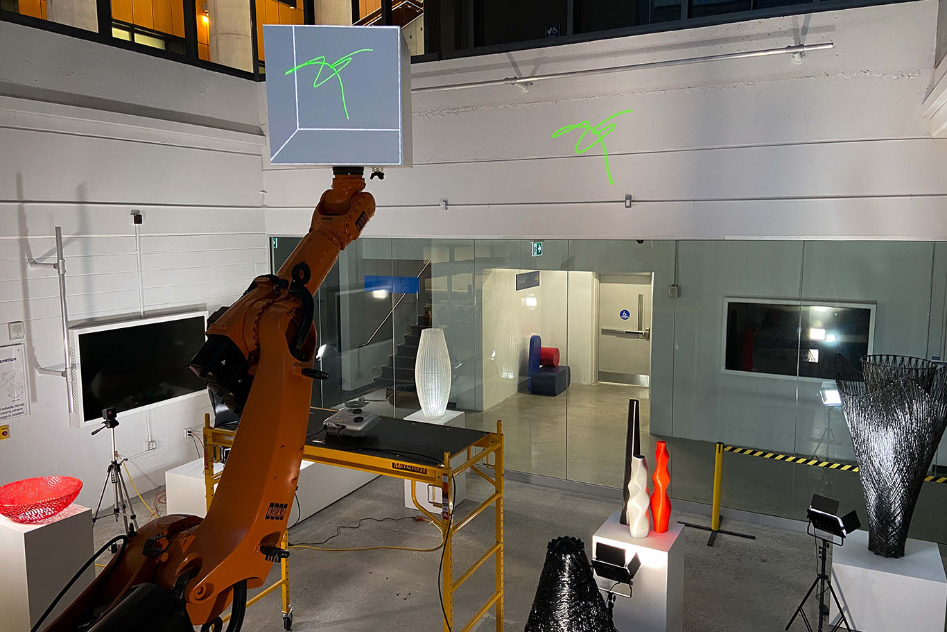 A large orange robotic arm holds up a white cube, and there is a green squiggly line being projected onto the cube's surface.